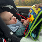 Kisbaba az autósülésben elmélyülten játszik a kék KalandTáska matatókönyvvel utazás közben.