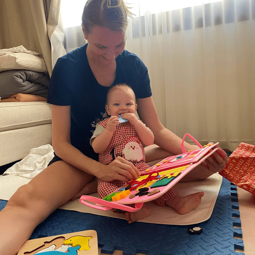 Édesanya és kisbabája együtt fedezik fel a pink KalandTáska interaktív elemeit a szőnyegen ülve.